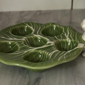 Puede incluir: Un plato de cerámica verde para servir huevos con forma de hoja de col, con seis huecos para huevos. Dos figuras de conejos de cerámica blanca flanquean el plato. El plato tiene vetas blancas.