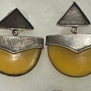May include: A pair of drop earrings featuring a triangular top piece connected to a semi-circular bottom section. The metal components have a textured silver-toned finish, while the lower half consists of a smooth, opaque yellow stone element.