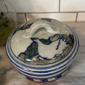 May include: A round ceramic lidded dish with a white and blue design. The lid features a handle and a painted bird motif. The dish has blue stripes around the base and a speckled finish.