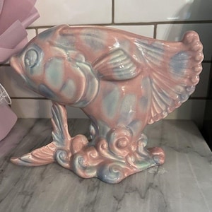 Puede incluir: Una figura de pez de cerámica en tonos rosa y azul. El pez tiene aletas detalladas y una base decorativa con patrones arremolinados en forma de nube. La figura se exhibe sobre una superficie de mármol.