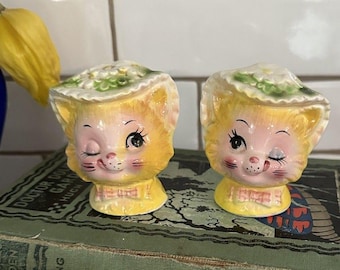 Puede incluir: Un par de saleros y pimenteros de cerámica con forma de gato, con pelaje amarillo, mejillas rosadas y gorros blancos con detalles florales verdes. Cada gato tiene un ojo guiñando, una pequeña nariz rosa y un lazo rosa en la base.
