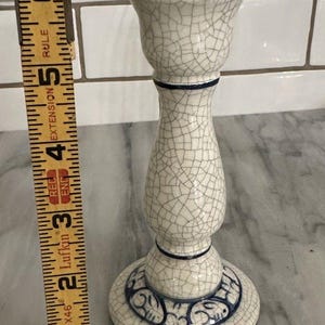 May include: A white ceramic candlestick with a crackle glaze finish. The candlestick has a round base with a blue decorative border, a slender stem, and a flared top. It measures approximately 5 inches tall.