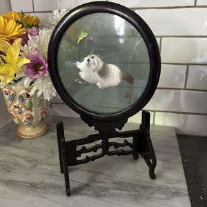 May include: A round, framed artwork featuring an embroidered cat gazing at a butterfly. The cat is white and gray, with blue eyes. The artwork is set in a dark wooden frame and displayed on a decorative stand.