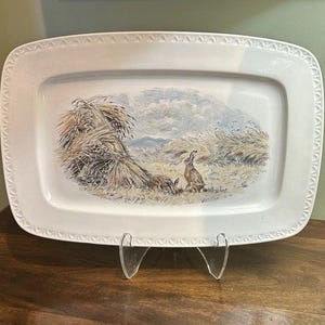 May include: A rectangular white ceramic serving platter featuring a decorative embossed border and a central illustration of two brown hares sitting in a field of tall golden wheat under a cloudy sky. The platter is displayed on a clear stand.