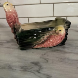 Op de afbeelding: Een keramische plantenbak met een rechthoekige vorm, met twee roze en gele parkietfiguren. De plantenbak heeft een donkergroene en zwarte basis, met een licht interieur. De parkieten zitten aan de zijkanten van de plantenbak.