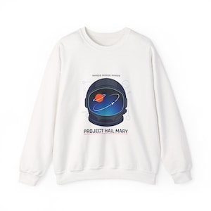 Könnte beinhalten: Weißes Sweatshirt mit einer Grafik eines Astronautenhelms, eines Planeten und Wirbellinien. Der Text "AMAZE AMAZE AMAZE" befindet sich über dem Helm, und "PROJECT HAIL MARY" steht darunter.