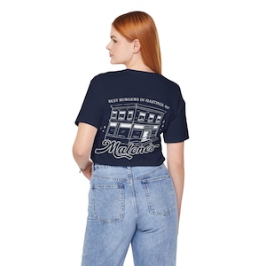 Puede incluir: Una camiseta azul marino de manga corta con un gráfico de líneas blancas en la espalda. El diseño muestra un edificio con el texto "BEST BURGERS IN HASTINGS, MA" encima y la palabra "Malone's" en letra cursiva debajo del edificio.