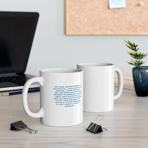 May include: Two white ceramic mugs with a curved handle. One mug has blue text that reads, "YOU THINK THIS HAS NOTHING TO DO WITH YOU... (THAT) LUMPY BLUE SWEATER..." The other mug is plain white.