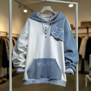 Pode incluir: Um moletom com capuz em blocos de cores branco e azul claro com um gráfico de gato na manga e um bolso frontal. O moletom com capuz tem um capuz com cordão e mangas compridas. Texto japonês impresso no bolso frontal.