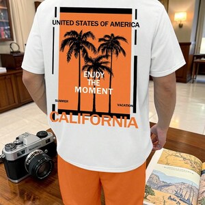 May include: White t-shirt with an orange graphic featuring palm trees and the text "Enjoy the Moment" and "California". The shirt is paired with orange shorts. A vintage camera sits on a wooden surface.