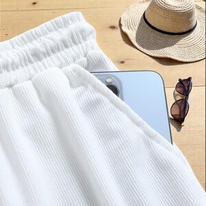 May include: Close-up of white textured shorts with a drawstring waistband. A straw sun hat with a navy blue band and sunglasses rest on a wooden surface. A smartphone is partially visible in the shorts pocket.