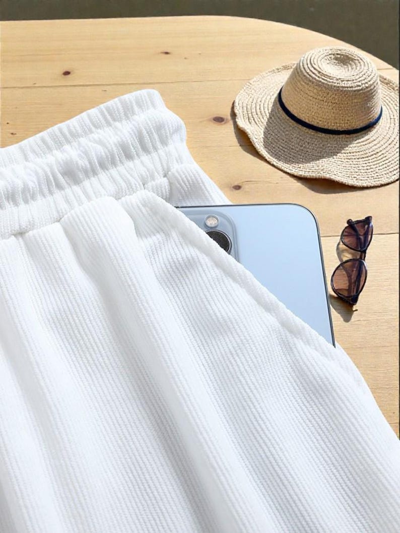 May include: Close-up of white textured shorts with a drawstring waistband. A straw sun hat with a navy blue band and sunglasses rest on a wooden surface. A smartphone is partially visible in the shorts pocket.
