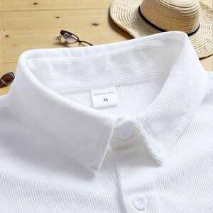 May include: Close-up of a white collared shirt with a button-down front. The shirt has a textured, ribbed fabric. A straw hat with a navy blue band and eyeglasses are in the background. The label reads "MADE IN CHINA" and "M".