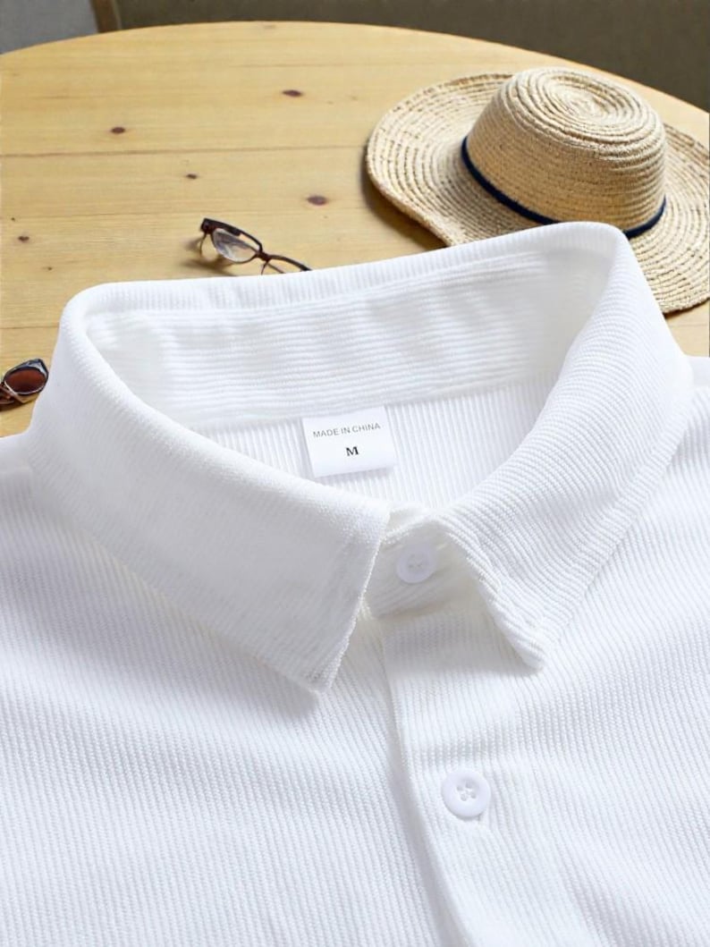May include: Close-up of a white collared shirt with a button-down front. The shirt has a textured, ribbed fabric. A straw hat with a navy blue band and eyeglasses are in the background. The label reads "MADE IN CHINA" and "M".