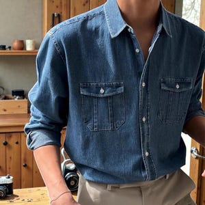 Puede incluir: Una camisa vaquera de lavado medio con dos bolsillos en el pecho y cierre de botones. La camisa tiene un cuello clásico y mangas largas. El modelo lleva la camisa con pantalones color caqui. La camisa es de estilo casual.