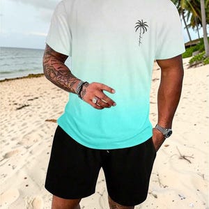 May include: White and turquoise ombre t-shirt with a palm tree graphic and black shorts. The t-shirt has a crew neck and short sleeves. The shorts are knee-length and have a drawstring waist. The outfit is worn on a sandy beach.