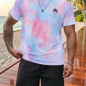 May include: A tie-dye t-shirt in shades of pink, blue, and white, featuring a small black palm tree graphic. The shirt is paired with black shorts. A silver necklace and bracelet are also visible.