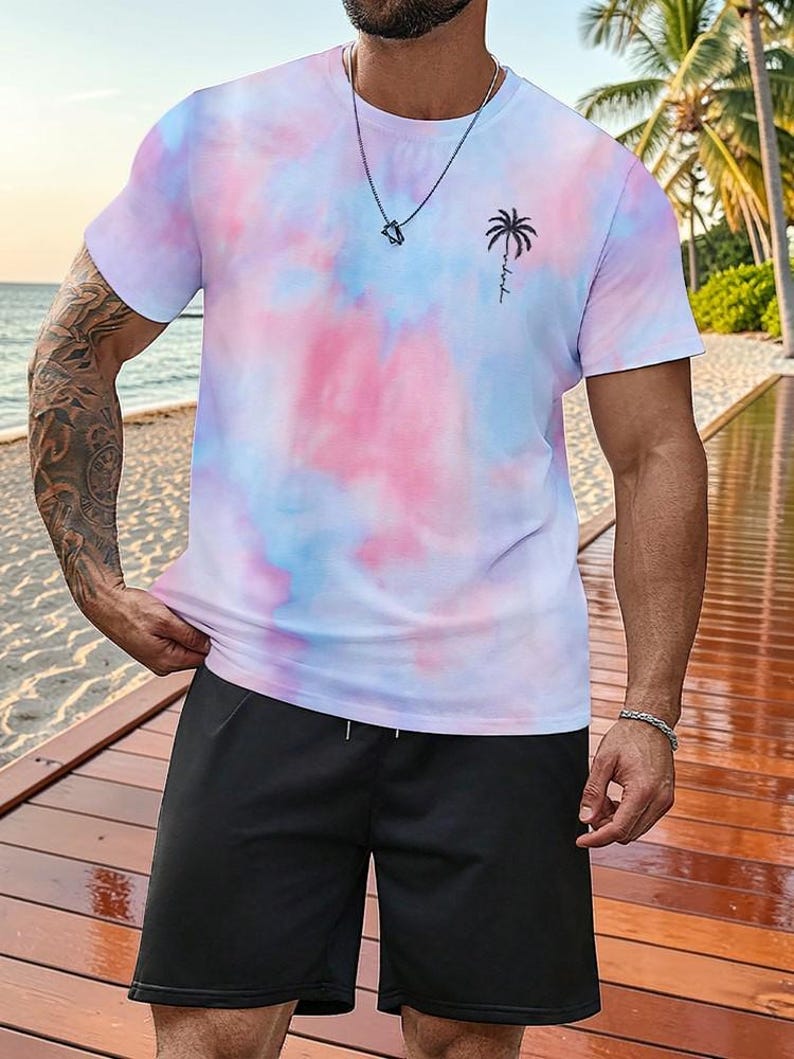 May include: A tie-dye t-shirt in shades of pink, blue, and white, featuring a small black palm tree graphic. The shirt is paired with black shorts. A silver necklace and bracelet are also visible.