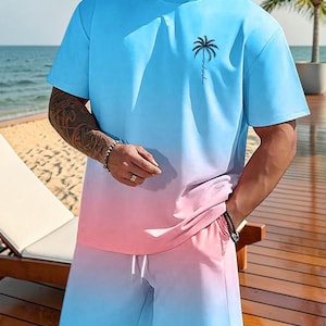 May include: A two-piece outfit featuring a blue and pink ombre design. The set includes a short-sleeved t-shirt with a palm tree graphic and matching shorts with a drawstring. The outfit is casual and suitable for warm weather.