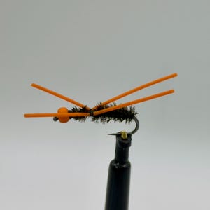 Könnte beinhalten: Nahaufnahme einer schwarzen Angel-Fliege mit orangefarbenen Akzenten. Die Fliege hat einen schwarzen Körper mit orangefarbenen Antennen und einer kleinen orangefarbenen Perle. Die Fliege ist an einem silbernen Haken befestigt.
