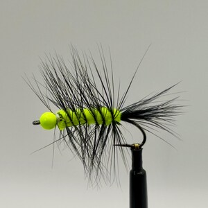 May include: A close-up of a fishing fly with a neon green body and a black, spiky hackle. The fly has a small, yellow bead head and is attached to a black hook.
