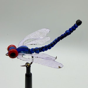 May include: A detailed dragonfly fishing lure with a blue body, clear wings, and red eyes. The dragonfly is mounted on a black stand, showcasing its intricate design and vibrant colors. This handcrafted lure is designed for fly fishing.