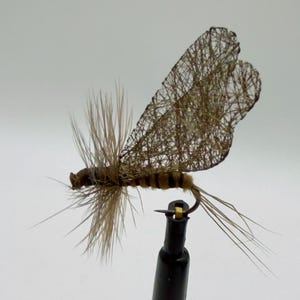 May include: A detailed close-up of a handmade fishing fly. The fly features a brown body with black stripes, tan feather-like appendages, and delicate, webbed wings. The fly is mounted on a black stand against a white background.