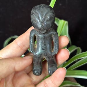 Könnte beinhalten: Eine kleine, dunkelgraue, anthropomorphe Steinfigur mit einer glatten, polierten Oberfläche. Die Figur hat einen abgerundeten Kopf mit geschnitzten Gesichtsvertiefungen, einen schlanken Torso mit offenen Seitenbereichen und zwei kurze, kräftige Beine.