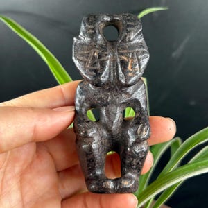 Könnte beinhalten: Ein dunkelgrauer, texturierter Steinanhänger, der als stilisierte anthropomorphe Figur geschnitzt ist. Das Stück verfügt über ein kreisförmiges Aufhängeloch an der Oberseite, zwei markante Gesichtszüge und offene Zwischenräume zwischen Armen und Beinen.