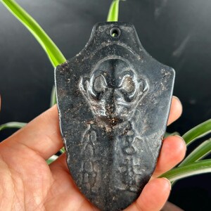 Könnte beinhalten: Ein schildförmiger Anhänger aus schwarzem Stein mit einer geschnitzten zentralen Figur mit großen, runden Augen und detaillierten Gesichtszügen, begleitet von vertikalen Reihen eingravierter Symbole und einem kleinen Loch oben.