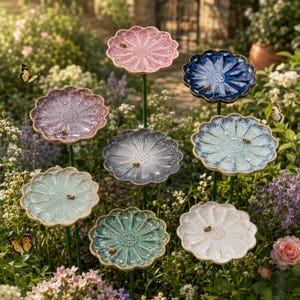 Könnte beinhalten: Eine Sammlung von acht blumenförmigen Vogeltränken aus Keramik auf dünnen Metallstäben. Jede Schale hat eine einzigartige glasierte Farbe, darunter Rosa, Dunkelblau, Violett, Grau, Hellblau, Türkis und Weiß, mit gewelltem Rand.