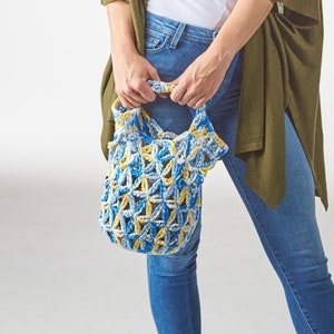 May include: A woman wearing a white tank top, blue jeans, and an olive green cardigan holds a blue, yellow, and white crocheted tote bag. The bag has a geometric pattern and two handles. The woman is smiling.