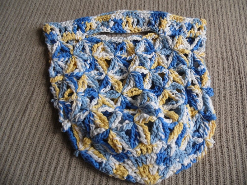 May include: A handmade crochet bag in a floral pattern. The bag features a combination of blue, white, and yellow yarn. The open weave design includes a handle at the top. The bag is likely used for carrying items.