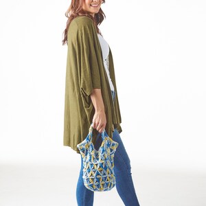 May include: A woman wearing a green cardigan, white top, and blue jeans, holding a blue and yellow crocheted tote bag. The bag has a geometric pattern. She is wearing black sandals. The background is white.