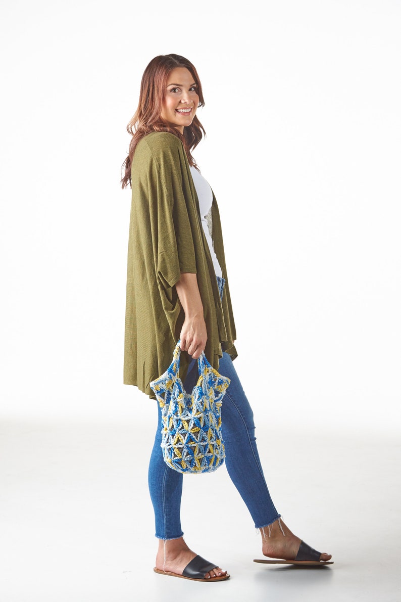 May include: A woman wearing a green cardigan, white top, and blue jeans, holding a blue and yellow crocheted tote bag. The bag has a geometric pattern. She is wearing black sandals. The background is white.