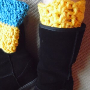 May include: A pair of black boots with yellow and blue crochet boot cuffs. The crochet cuffs are visible above the boots, adding a pop of color. The boots appear to be made of a soft material.