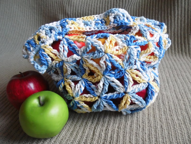 May include: A handmade crochet bag in blue, white, and yellow floral pattern, holding red and yellow apples. A red apple and a green apple are next to the bag. The bag has a handle and is open at the top.