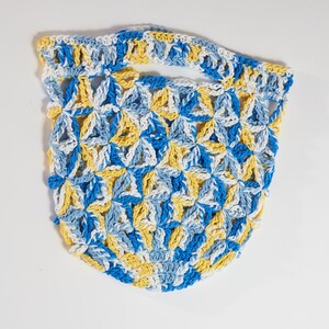 May include: A handmade crochet bag in a floral pattern. The bag features a handle and is made with blue, yellow, and white yarn. The bag is a light, airy design, perfect for carrying small items.