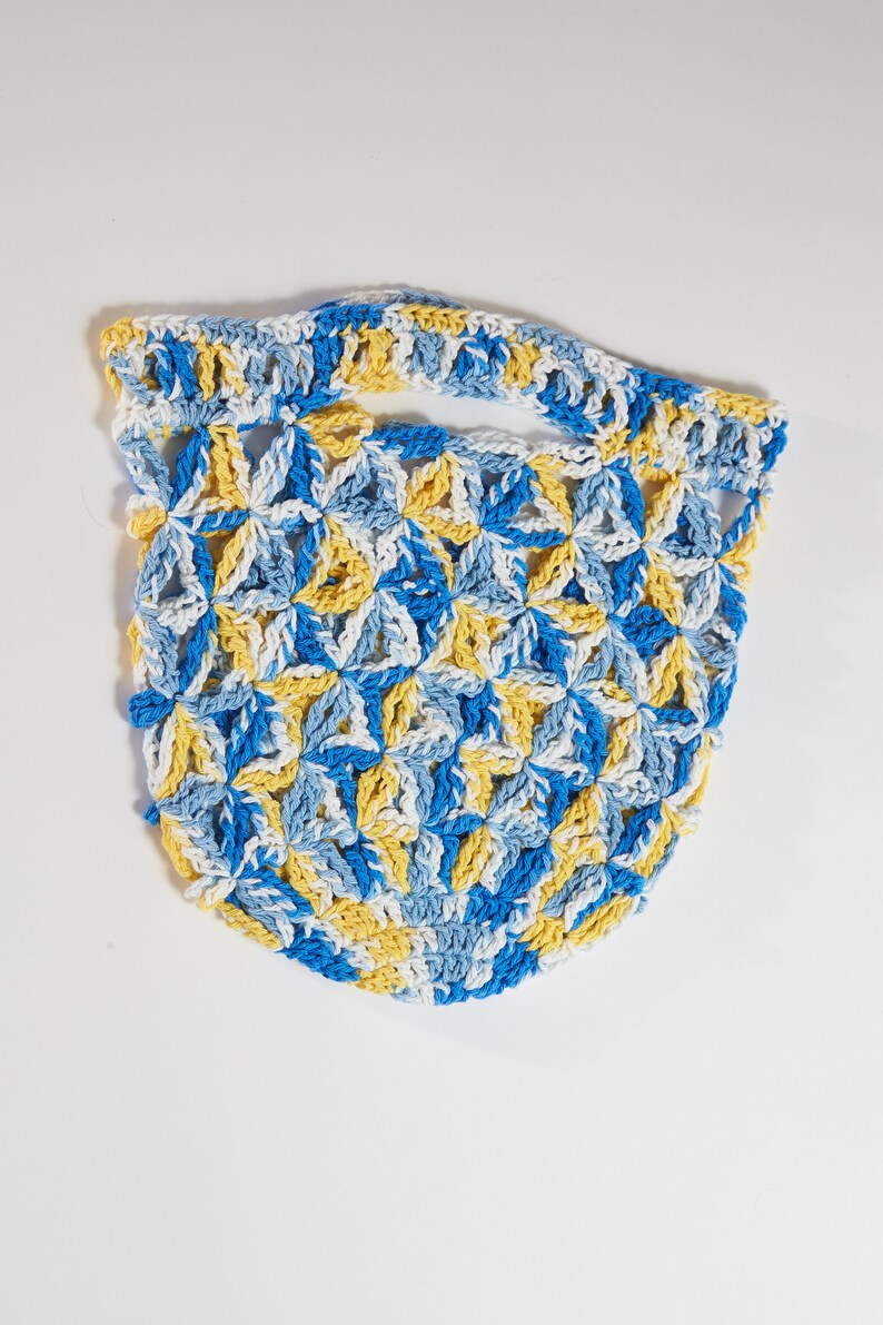 May include: A handmade crochet bag in a floral pattern. The bag features a handle and is made with blue, yellow, and white yarn. The bag is a light, airy design, perfect for carrying small items.