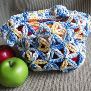 May include: A handmade crochet bag in shades of blue, white, and yellow, filled with red and yellow apples. The bag has a floral pattern and a handle. A red apple and a green apple sit beside the bag.
