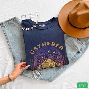 May include: Navy blue t-shirt with the word "GATHERER" in gold lettering. The shirt features a stained glass design with stars and a sun. A brown hat and light blue jeans are also in the image.