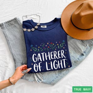 May include: A navy blue sweatshirt with the words "GATHERER OF LIGHT" in white script, decorated with a floral design. The sweatshirt is paired with light-wash jeans and a brown hat. A hand with a ring and bracelet is visible.