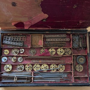 May include: An open, antique wooden box with a red velvet interior, filled with metal construction toy parts. The box contains various wheels, rods, and plates, suggesting a vintage building set.