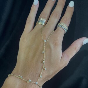 Könnte beinhalten: Ein goldfarbenes Handkettenarmband mit herabhängenden, klaren Edelsteinen. Das Armband verbindet sich mit einem Ring und verfügt über mehrere kleine, klare Edelsteine. Mehrere Ringe sind ebenfalls an den Fingern zu sehen.