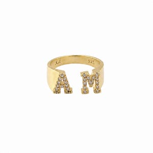 Könnte beinhalten: Ein goldfarbener offener Ring mit den Buchstaben A und M als Blickfang. Die Buchstaben sind mit kleinen, klaren, funkelnden Steinen besetzt. Die Innenseite des Rings ist deutlich mit der Prägung 925 versehen, was auf Sterlingsilber hinweist.
