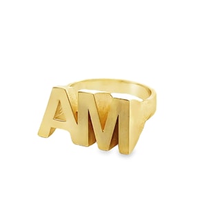 May include: A gold-colored ring with the letters "AM" prominently displayed. The ring has a thick band and the letters are raised and blocky. The ring is set against a white background.