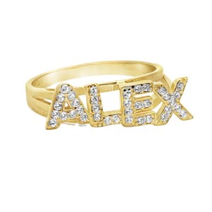 Könnte beinhalten: Ein goldfarbener Ring mit dem Namen "ALEX", der in funkelnden, klaren Edelsteinen geschrieben ist. Der Ring hat ein schlichtes, glattes Design, während die Buchstaben mit den Edelsteinen besetzt sind, was ein personalisiertes und auffälliges Accessoire ergibt.