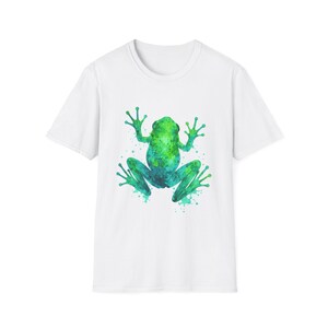 May include: White t-shirt featuring a watercolor frog design. The frog is depicted in shades of green and blue, with a splattered paint effect. The frog is centered on the shirt.