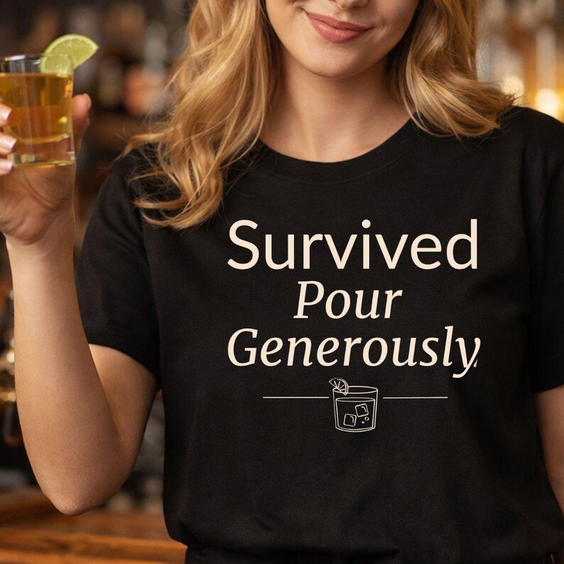 May include: Black t-shirt with the white text "Survived Pour Generously" and a cocktail graphic. The shirt is a crew neck style and appears to be made of a soft material. A person is holding a shot of liquor with a lime wedge.