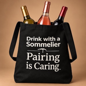 May include: A black tote bag featuring the white printed text Drink with a Sommelier, a graphic of a corkscrew, and the text Pairing is Caring. Three wine bottles with gold, pink, and dark red foil capsules are tucked inside the bag.
